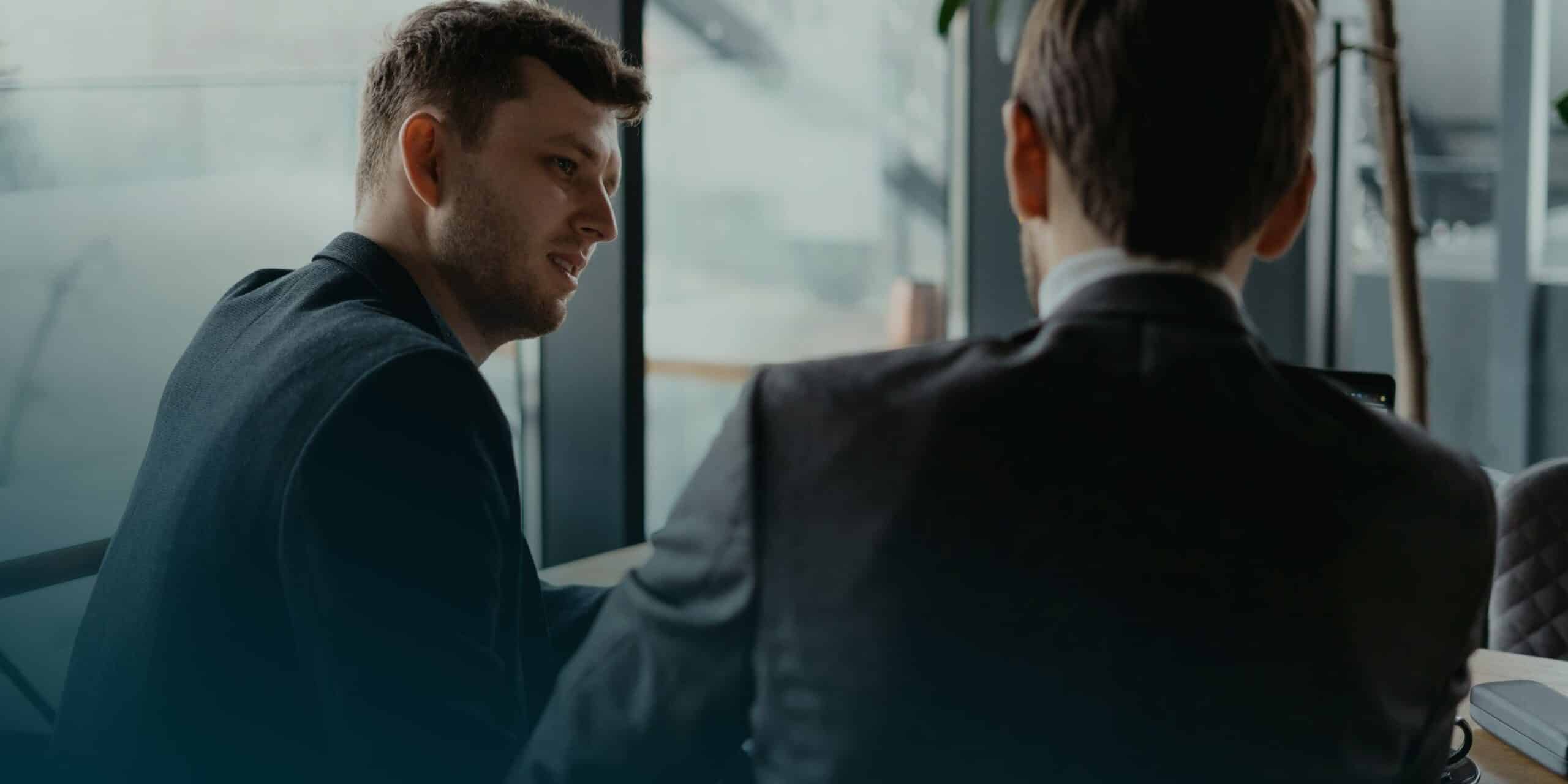 Two men in suits are engaged in conversation at a modern office with large windows, suggesting a professional setting.