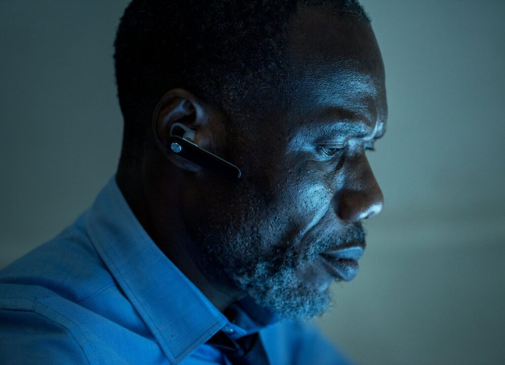 Man in a blue shirt using a headset, illuminated by a soft blue light, focusing intently in a dimly lit setting.