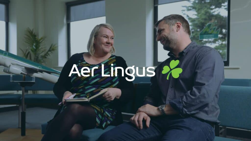 Two people sitting and smiling at each other in an office setting, with the Aer Lingus logo displayed over them.