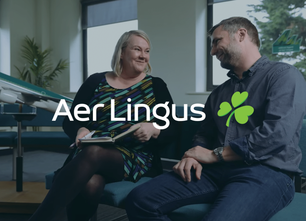Two people sitting and smiling at each other in an office setting with the Aer Lingus logo and shamrock symbol displayed across the image.