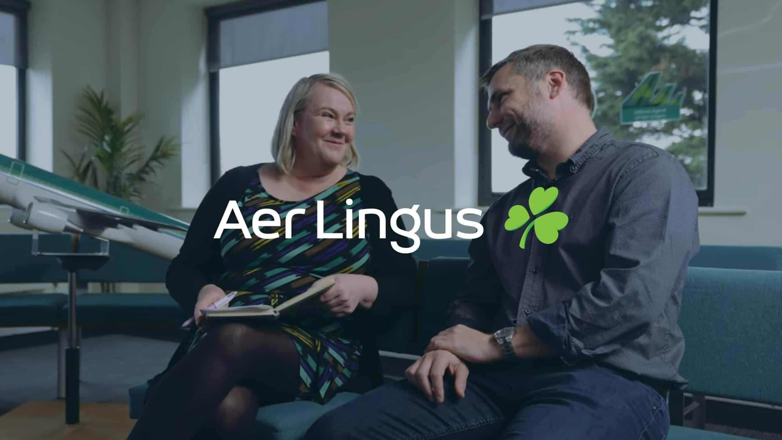 Two people sitting and smiling at each other in an office setting, with the Aer Lingus logo displayed over them.