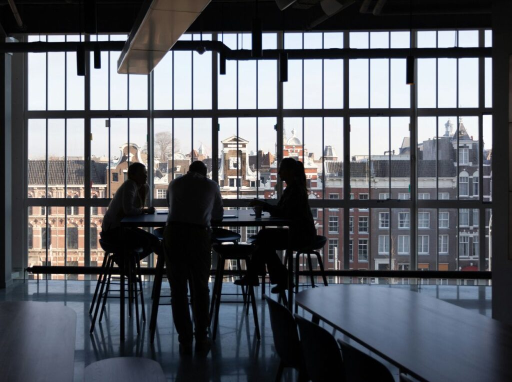 Three people sit at a table in front of large windows overlooking city buildings, silhouetted by daylight.
