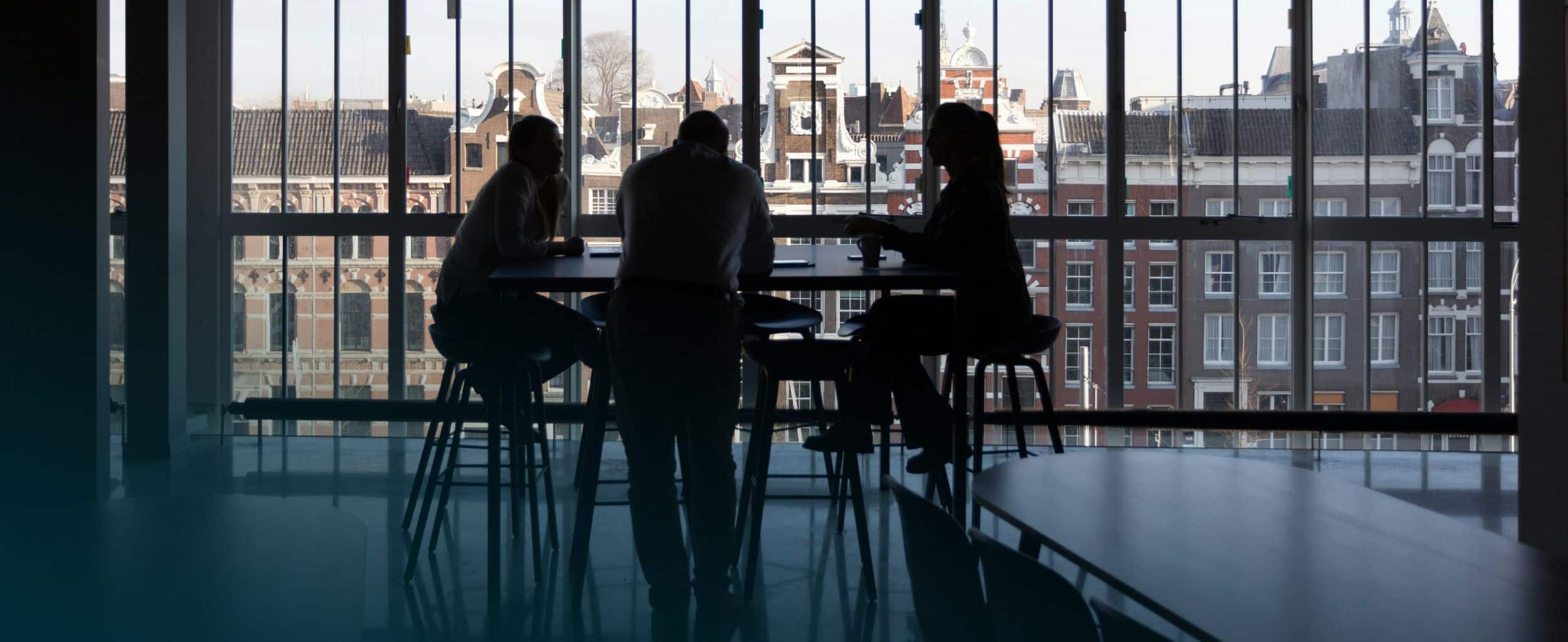 Three people sit at a round table silhouetted against large windows, overlooking a cityscape.