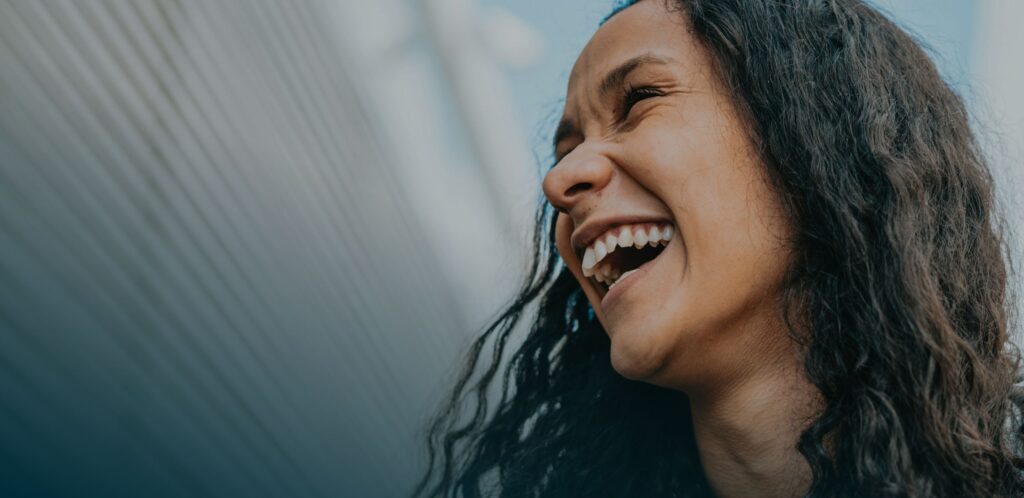 A woman with long, curly hair is smiling and looking upward, set against a blurred outdoor background.