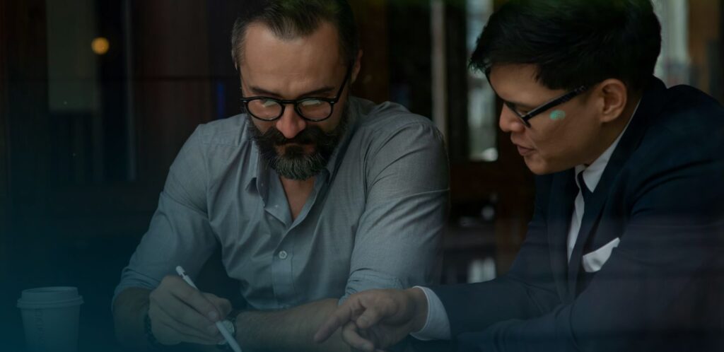 Two men sitting at a table discussing documents, one pointing at the paper, both wearing glasses.