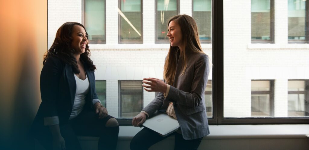 Two women sit on a windowsill in an office setting, engaged in a friendly conversation, with one holding a notebook.