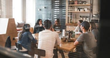A group of people sitting around a wooden table in a casual meeting room, discussing collaboratively.