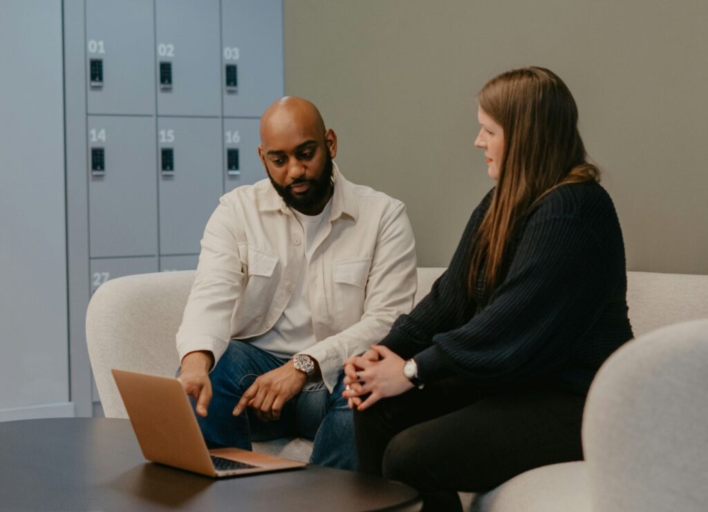 Two people seated on a couch, discussing something on a laptop, with lockers in the background.