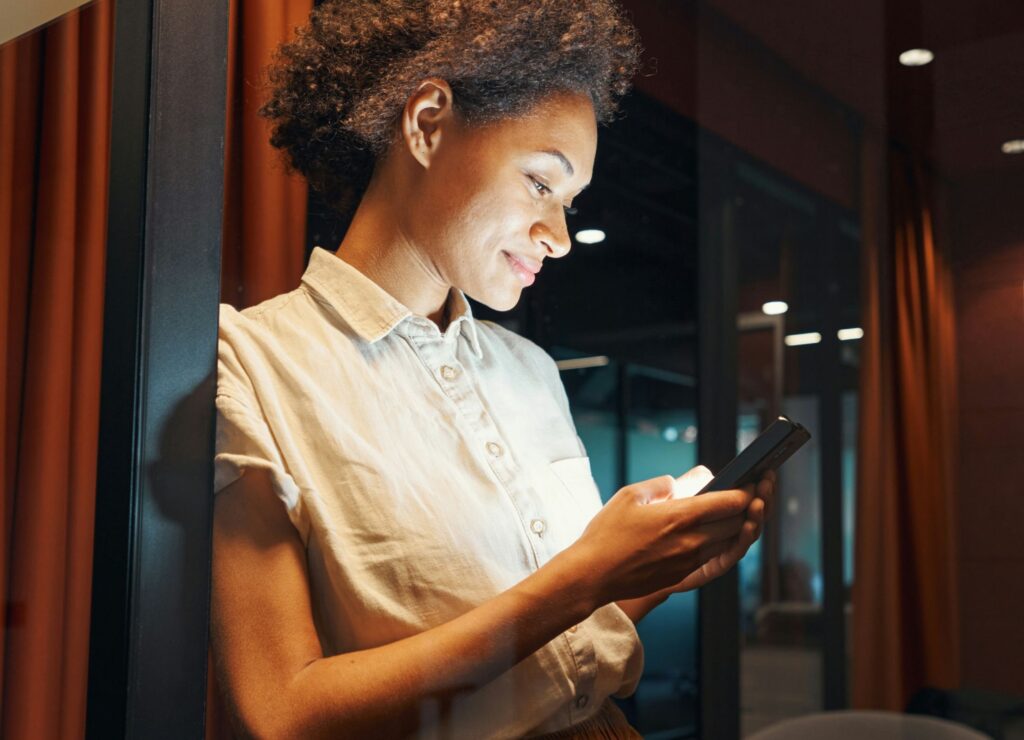 A woman stands indoors, smiling while looking at her smartphone, with warm lighting in the background.