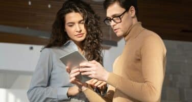 Two people stand together, examining a tablet. One wears a light blue shirt, the other a beige sweater. They appear focused and are in a well-lit, modern interior space.