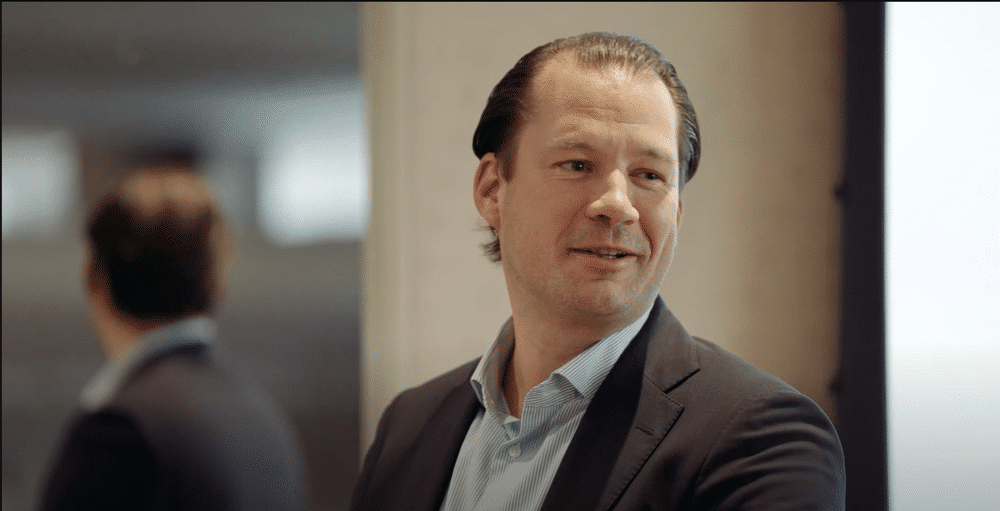 A person in a suit and blue shirt smiles slightly and appears to be engaged in conversation. A mirror in the background shows the person’s reflection.