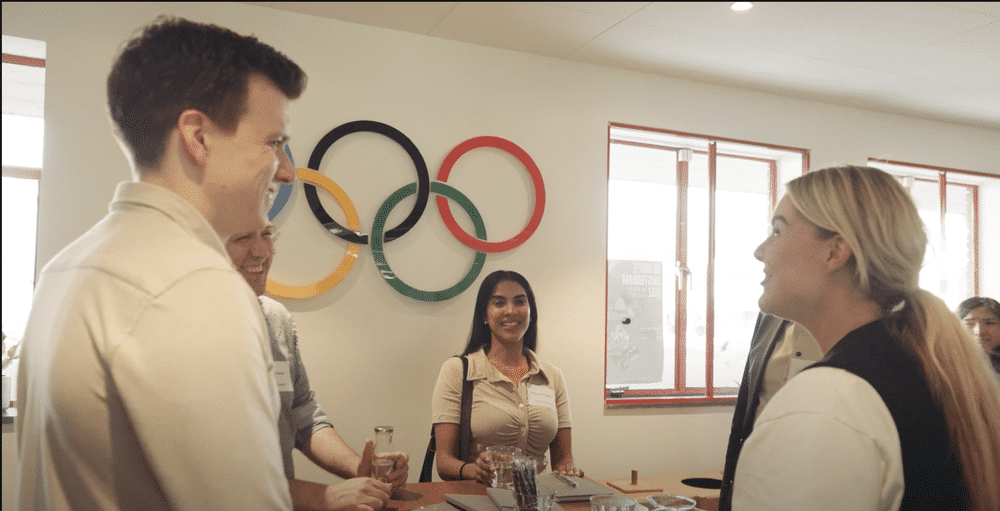 A group of people in a meeting room are smiling and conversing. Olympic rings are displayed on the wall in the background.