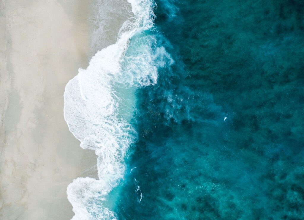 Aerial view of ocean waves crashing onto a sandy beach, with vibrant teal water contrasting the light sand.