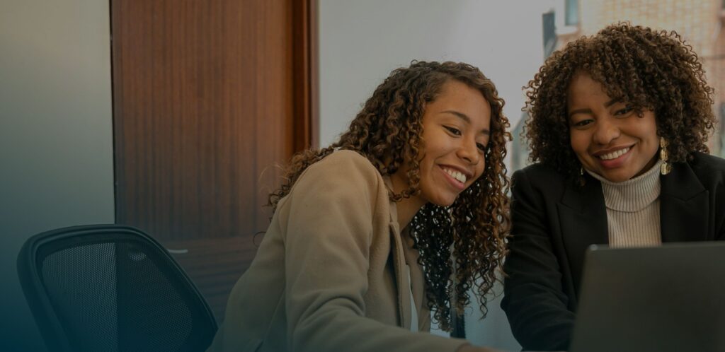 Two people with curly hair smiling and looking at a laptop screen in an office setting.