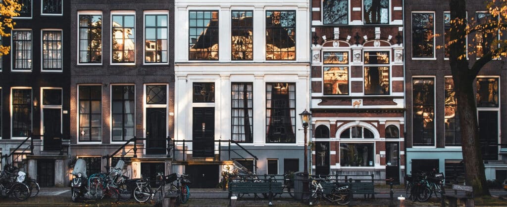 Row of traditional Amsterdam canal houses with large windows, bicycles parked along the street, and warm interior lights visible.