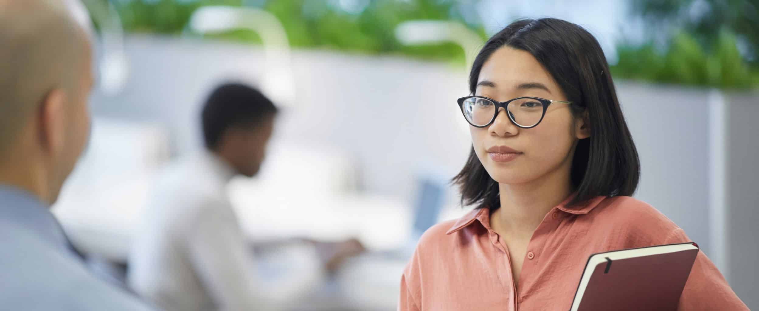 A woman with glasses holds a notebook, listening to a bald man in an office environment. Another man is blurred in the background, seated and working at a desk.