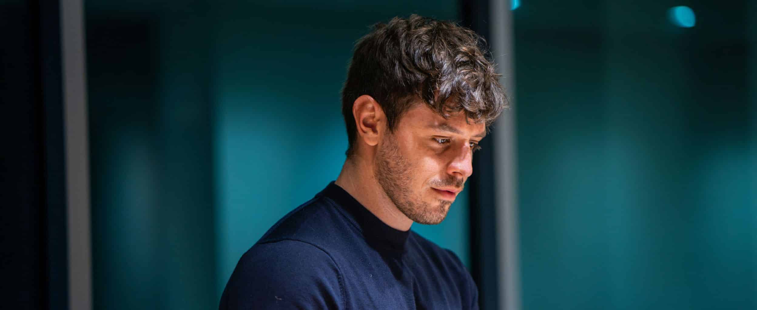 A man with short, wavy hair wearing a dark shirt gazes intently downward, set against a softly blurred, teal-colored background.