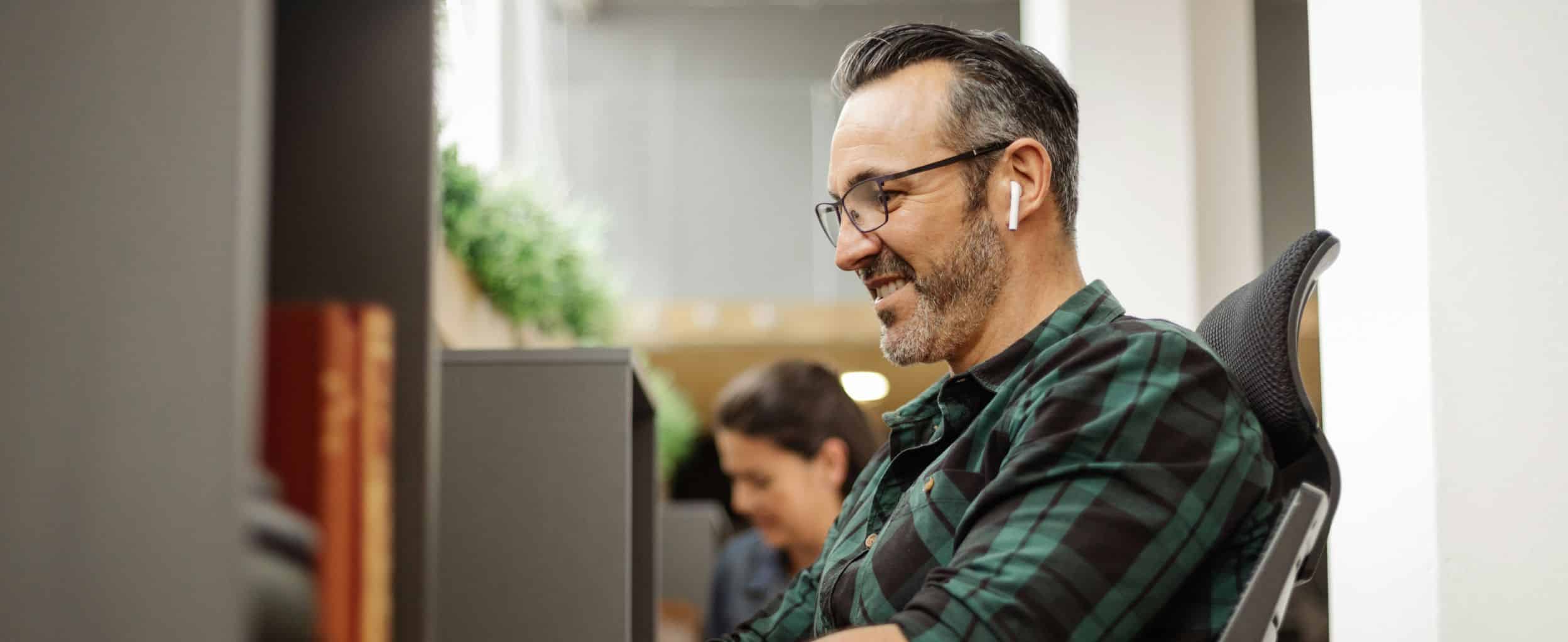 A man wearing wireless earbuds is smiling while looking at a computer screen in an office setting.