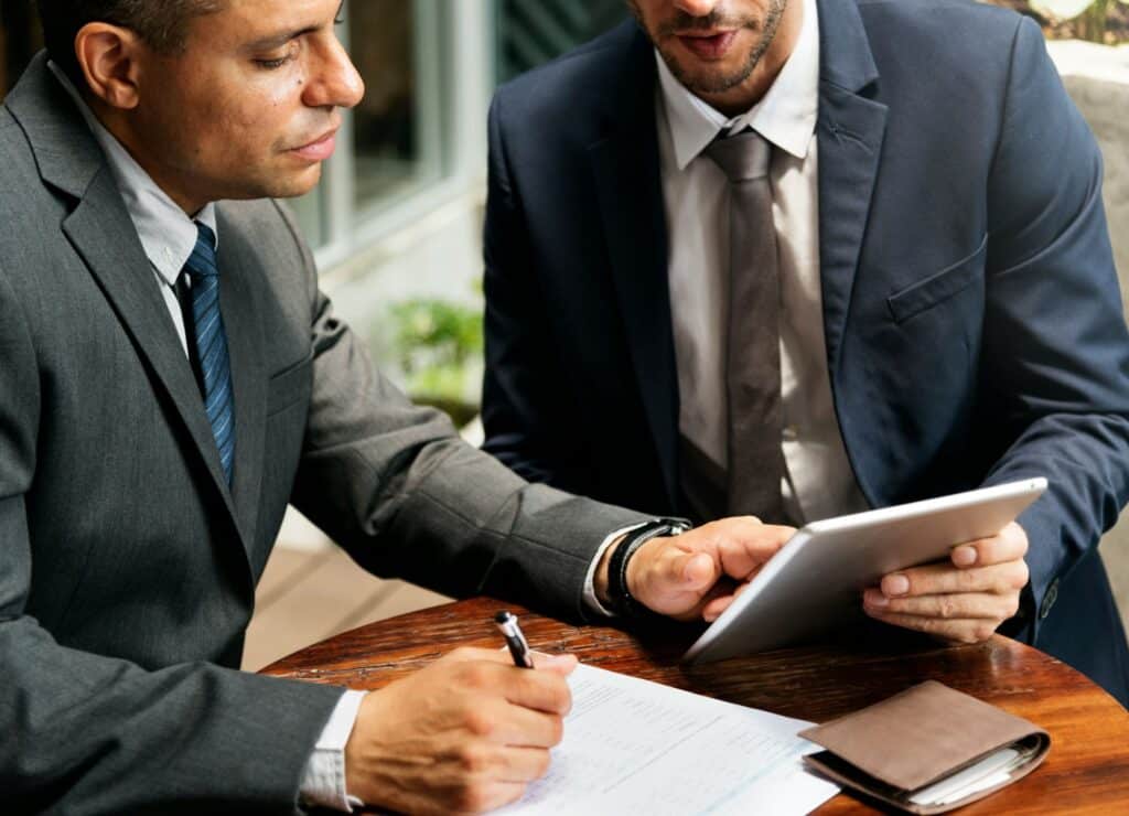 Two businessmen in suits are sitting at a table, discussing while looking at a tablet. One is taking notes on paper.
