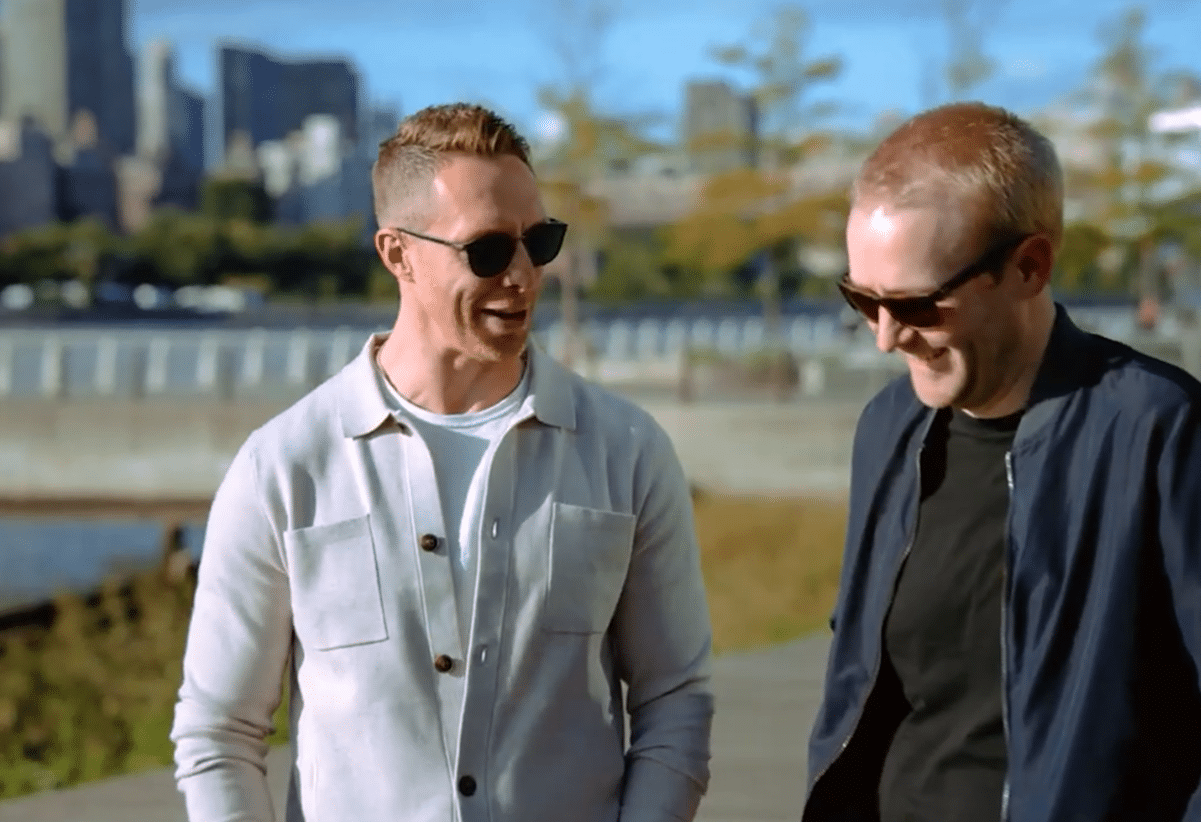 Two men wearing sunglasses are walking and laughing together outdoors, with a city skyline and river in the background.