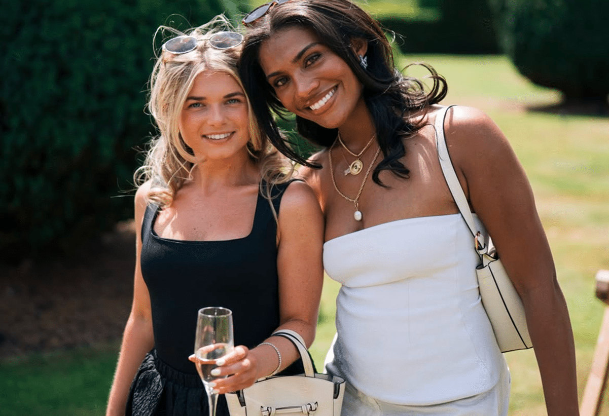 Two women smiling outdoors in a garden, dressed in stylish attire. One holds a glass of champagne, and both carry handbags.