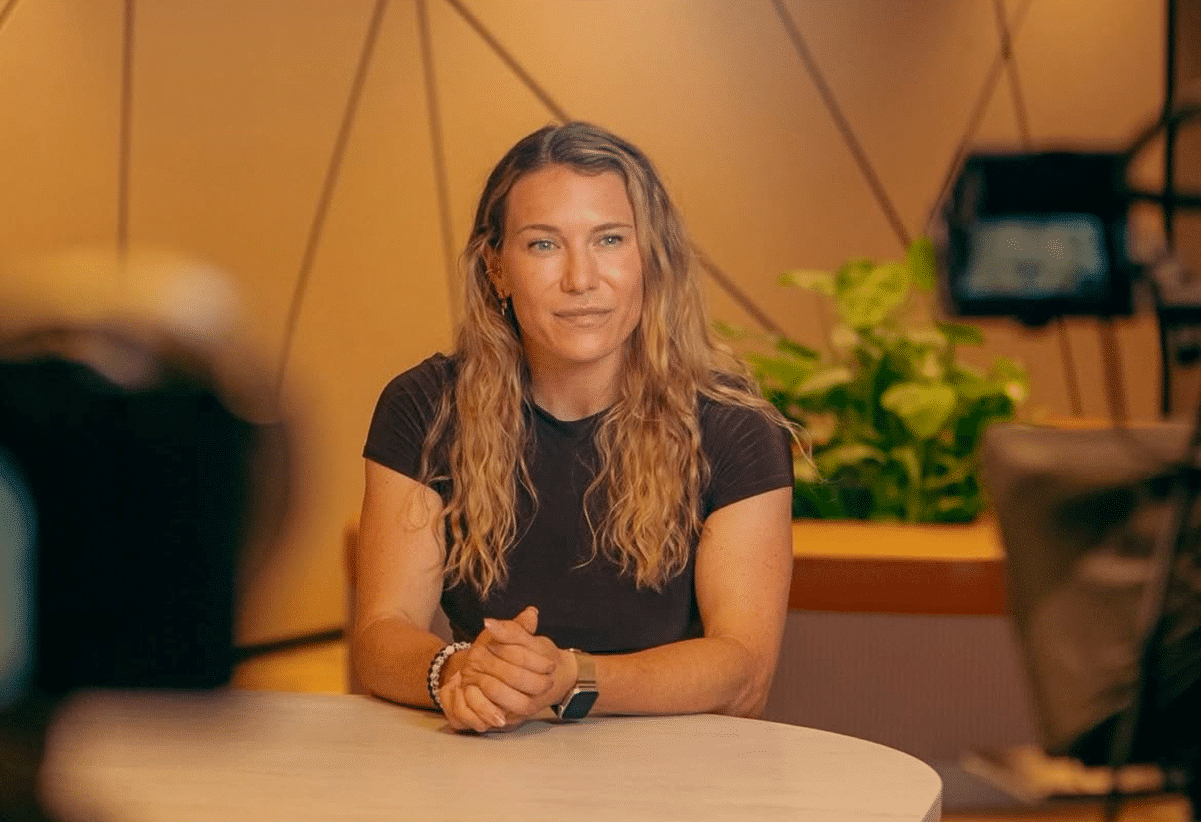 A person with long hair sits at a table, facing the camera, with a relaxed posture. The background includes geometric wall patterns and a plant, and there are camera setups visible around them.