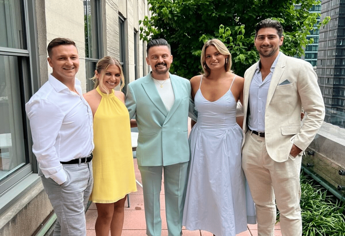 Five people dressed in summer formal attire stand smiling on a balcony with greenery and tall buildings in the background.