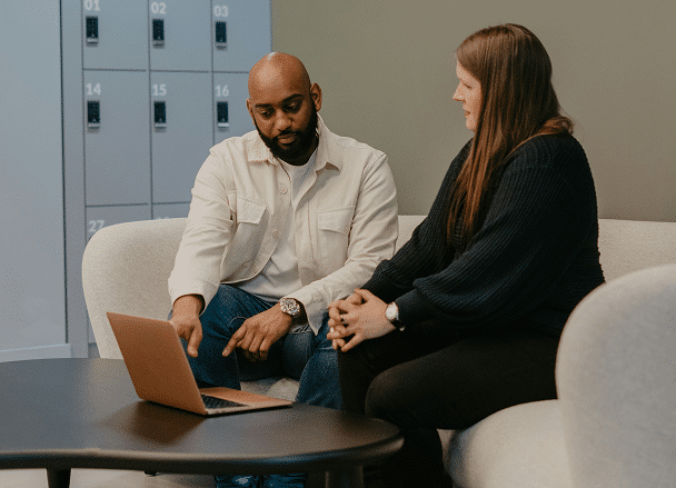 Two people sitting on a couch in a modern office, discussing something on a laptop.