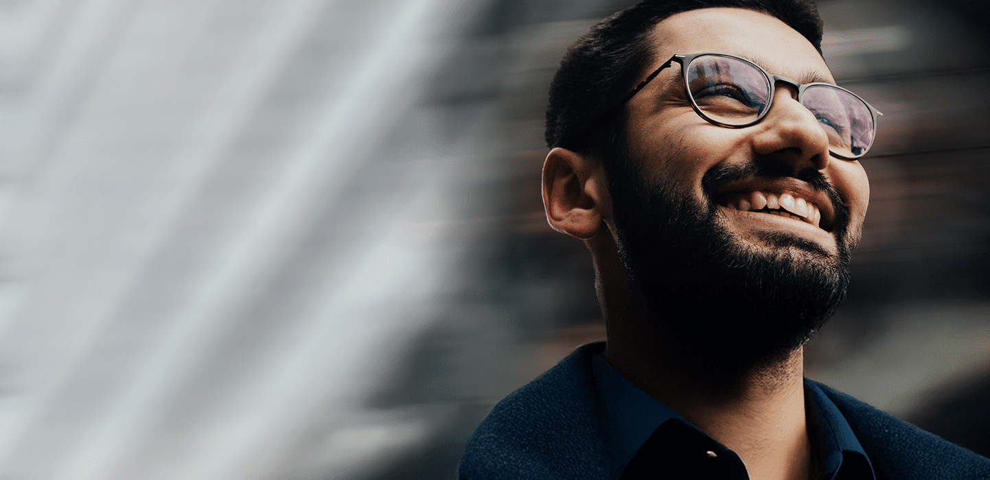 A man with a beard and glasses is smiling while looking upward. The background is blurred, featuring diagonal light streaks.