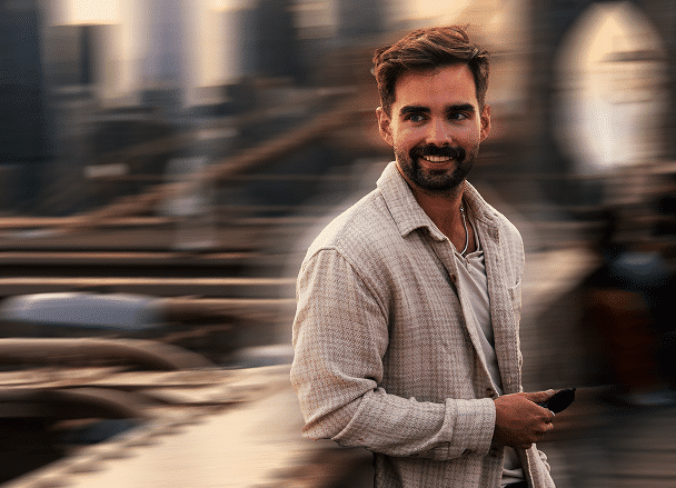 A man in a light jacket stands on a bridge smiling, with city buildings blurred in the background, creating a sense of motion and urban life.