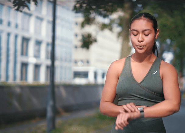 A woman in athletic wear checks her smartwatch while standing on an outdoor path lined with trees and buildings.