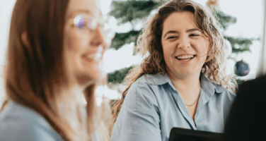 Two women smiling and talking indoors with a decorated Christmas tree in the background.