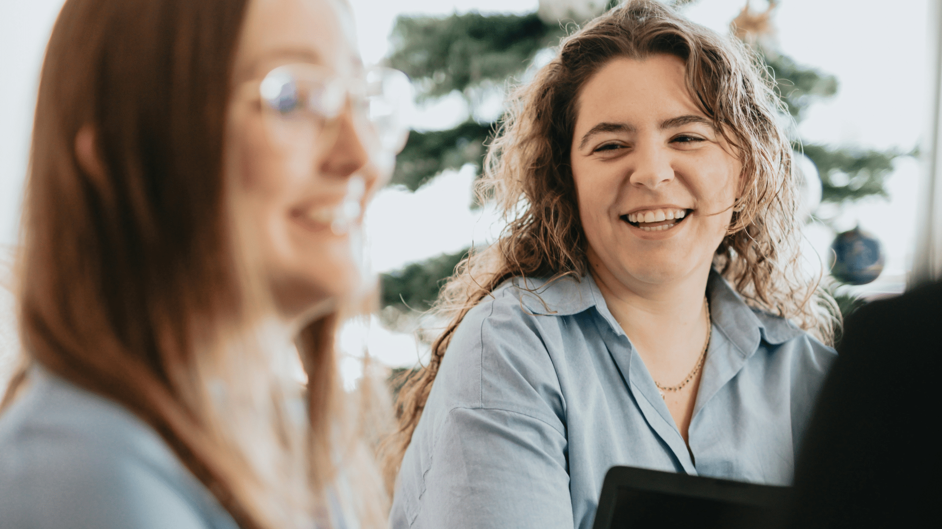 Two women smiling and talking indoors with a decorated Christmas tree in the background.
