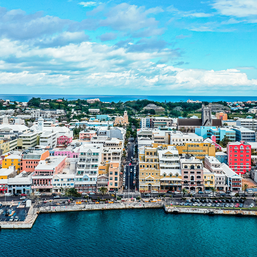 Aerial view of a coastal cityscape featuring colorful buildings along the waterfront, against a backdrop of lush greenery and a partly cloudy sky.