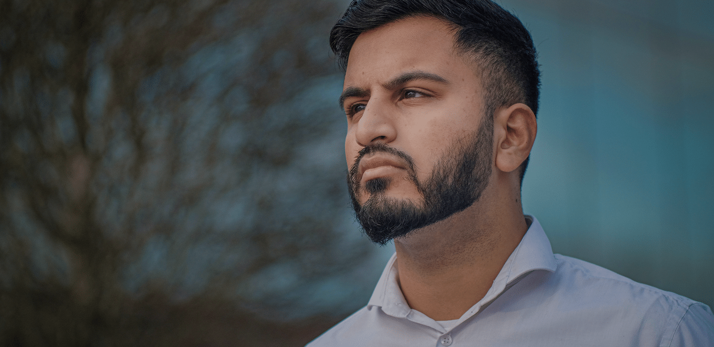 A man with a beard and a contemplative expression looks into the distance, with a blurred background of trees and a building.