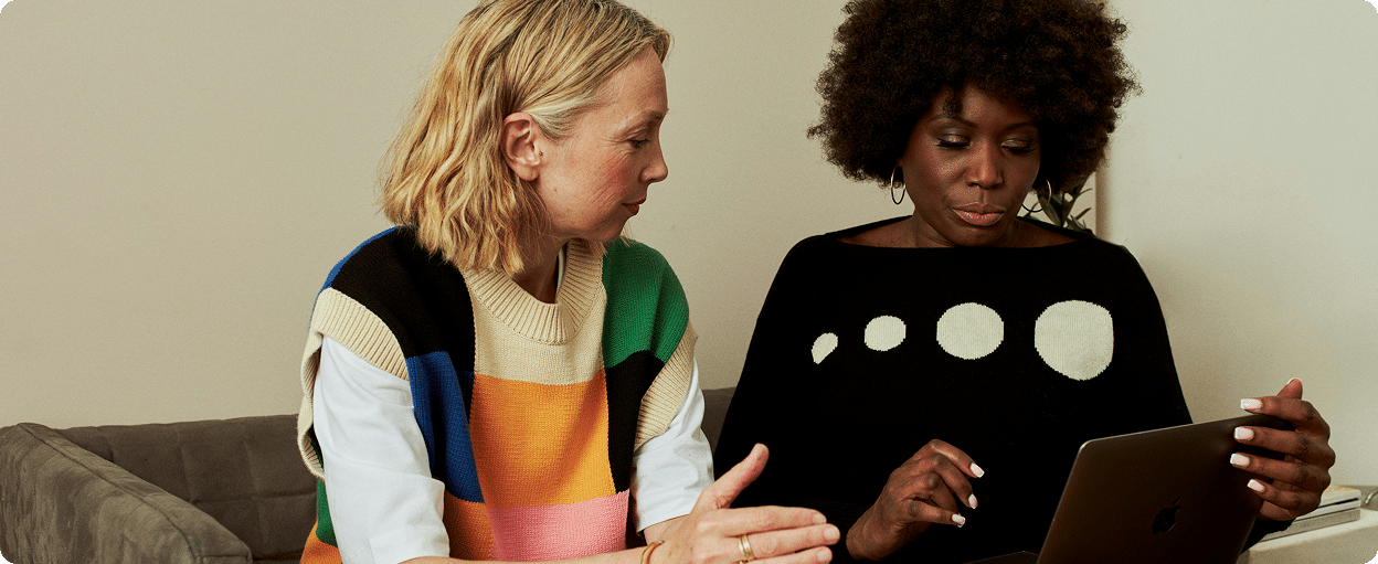 Two women sitting on a couch, engaged in a discussion while looking at a tablet.