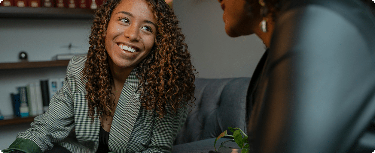 Two people sitting on a couch engaged in a conversation, with one person smiling warmly.