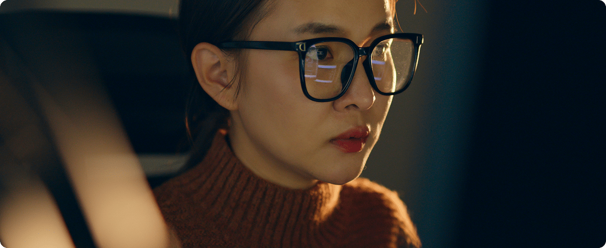 A woman wearing glasses and a brown sweater is focused on a computer screen, illuminated by its light.