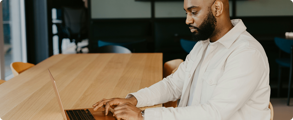 A man in a white shirt works intently on a laptop at a wooden table in a modern office setting.