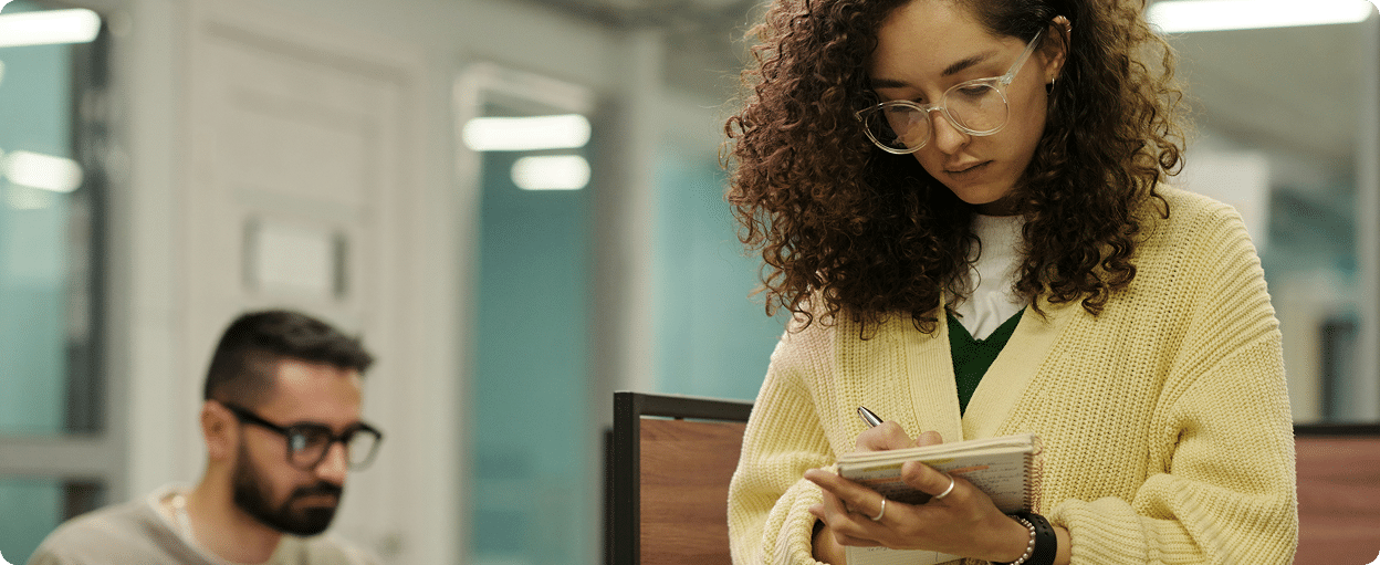 A woman in glasses and a yellow sweater writes in a notebook while standing in an office; a man works on a computer in the background.
