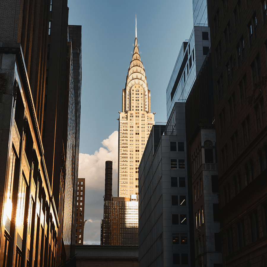The image shows the Chrysler Building towering between shadowed city skyscrapers, illuminated by the setting sun against a partly cloudy sky.