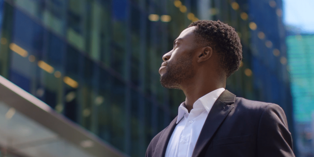 A confident professional in a suit looks upward, standing against a background of modern glass buildings, suggesting ambition and focus in an urban setting.