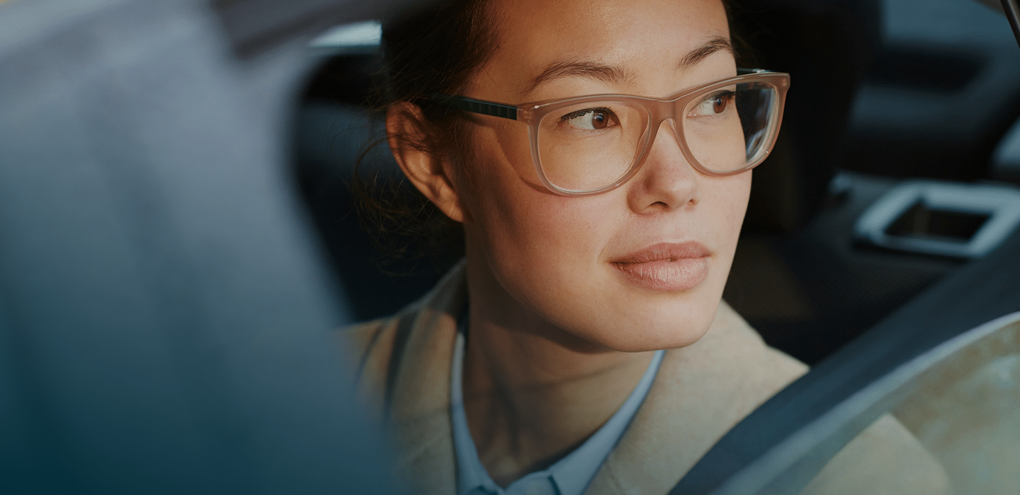 A person with glasses gazes thoughtfully out of a car window, sunlight softly illuminating their face.