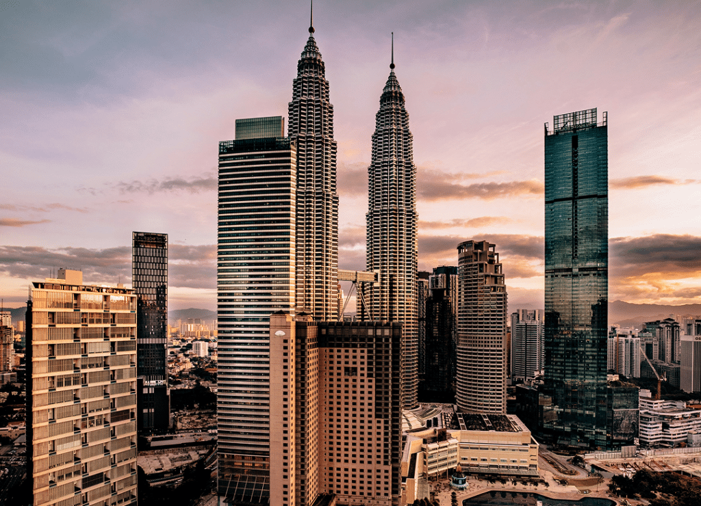 A city skyline at sunset featuring prominent skyscrapers, including two iconic twin towers, with a backdrop of a colorful sky.