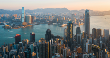 Aerial view of Hong Kong skyline at sunset, featuring tall skyscrapers, a bustling harbor, and distant mountains under a warm, golden sky.
