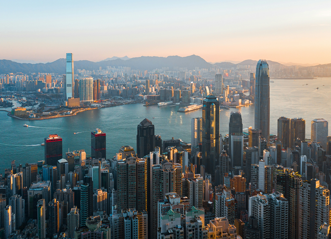 Aerial view of Hong Kong skyline at sunset, featuring tall skyscrapers, a bustling harbor, and distant mountains under a warm, golden sky.