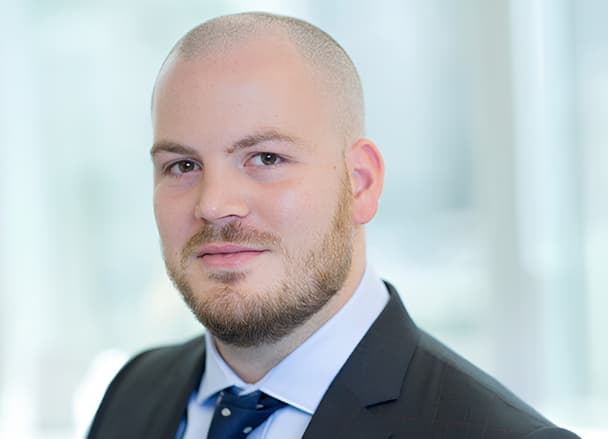 A professional man with a bald head and beard is wearing a suit and tie, standing against a blurred background.