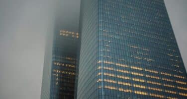 Two tall skyscrapers rise into a cloudy sky, with office lights glowing warmly in the windows.