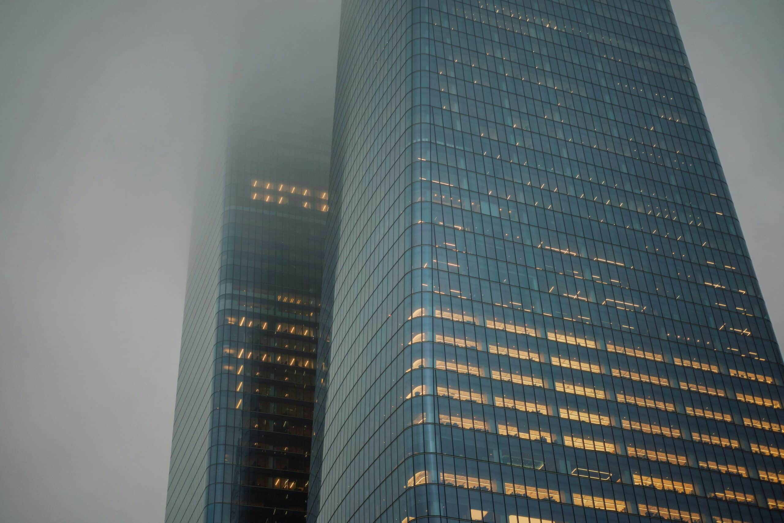 Two tall skyscrapers rise into a cloudy sky, with office lights glowing warmly in the windows.