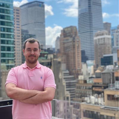 A person in a pink polo shirt stands confidently on a rooftop, with a city skyline featuring tall buildings and blue sky in the background.